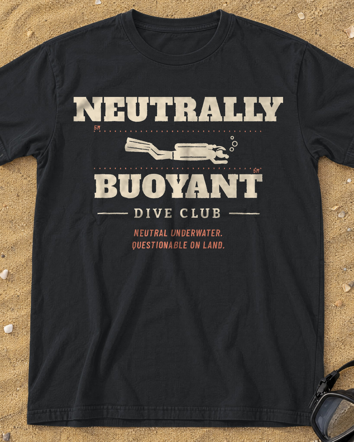 Black Neutrally Buoyant Dive Club tee with sand and coral scuba diver print, laid on a sandy beach