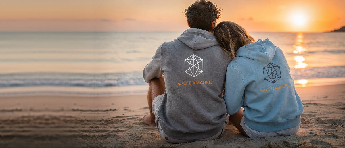 A couple in Salt Damaged hoodies sitting on the beach at sunset